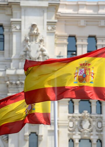 Primer plano de banderas de España ondeando al viento frente a un edificio emblemático de Madrid, simbolizando el impulso nacional al nuevo Plan de Derechos Culturales.