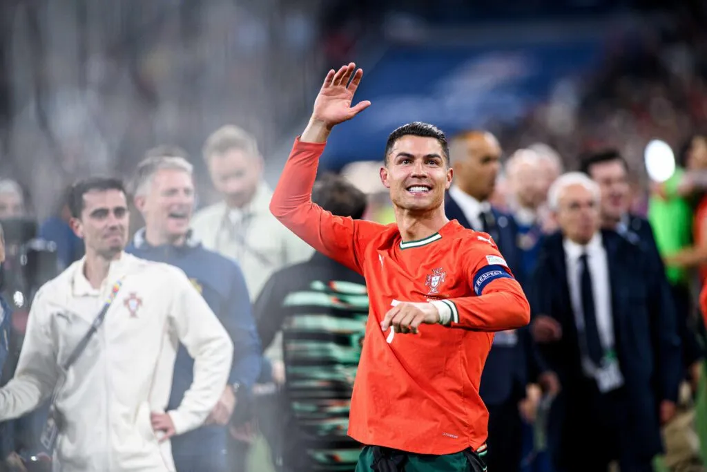 Cristiano Ronaldo, con uniforme rojo de la selección portuguesa, sonríe y saluda a la afición en un estadio lleno, rodeado de compañeros y personal técnico.
