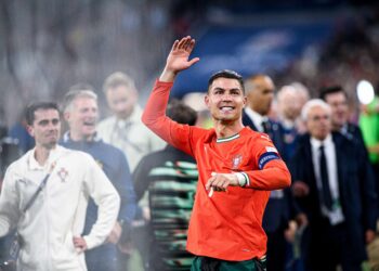 Cristiano Ronaldo, con uniforme rojo de la selección portuguesa, sonríe y saluda a la afición en un estadio lleno, rodeado de compañeros y personal técnico.