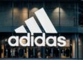 Vista frontal de una tienda Adidas con su logotipo iluminado, exhibiendo ropa deportiva en su interior.