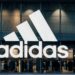 Vista frontal de una tienda Adidas con su logotipo iluminado, exhibiendo ropa deportiva en su interior.