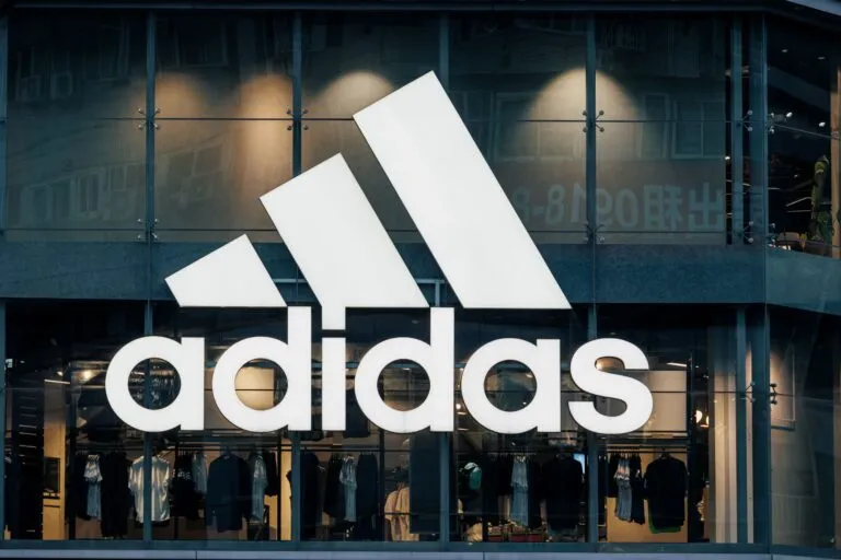 Vista frontal de una tienda Adidas con su logotipo iluminado, exhibiendo ropa deportiva en su interior.