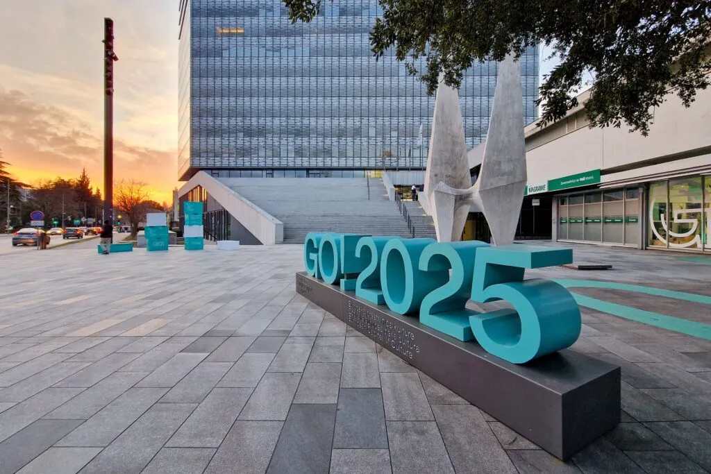 Escultura urbana con el logotipo “GO! 2025” en Gorizia, Italia, anunciando su designación como Capital Europea de la Cultura, con edificios modernos y atardecer de fondo.