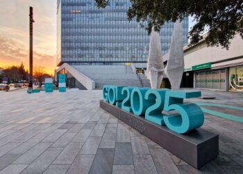 Escultura urbana con el logotipo “GO! 2025” en Gorizia, Italia, anunciando su designación como Capital Europea de la Cultura, con edificios modernos y atardecer de fondo.