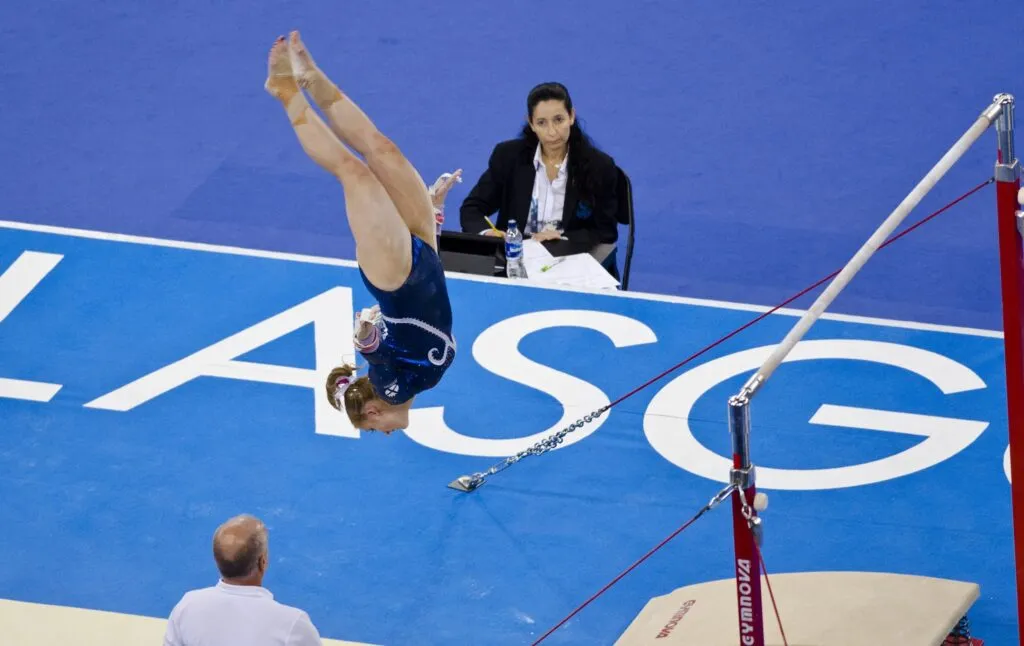 Atleta de gimnasia artística realiza un salto invertido en barras asimétricas bajo la supervisión de un juez y su entrenador, en un evento deportivo de alto nivel.