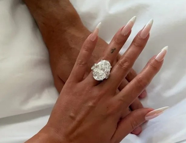 Primer plano de la mano de Georgina Rodríguez luciendo un gran anillo de diamante ovalado, entrelazada con la mano de Cristiano Ronaldo sobre sábanas blancas.