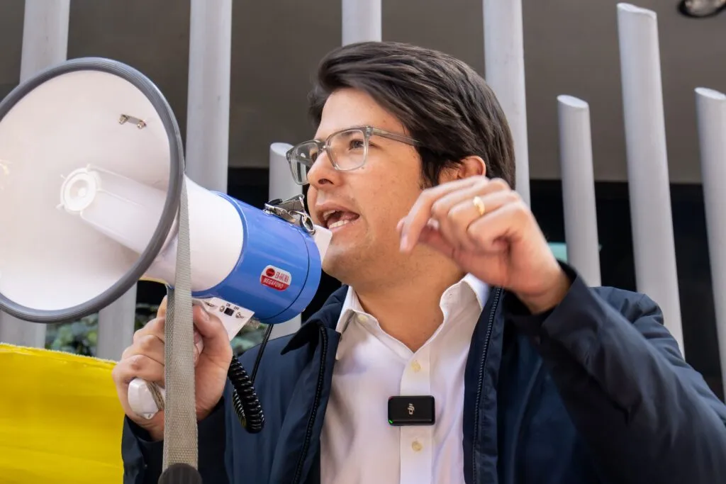 Miguel Uribe Turbay habla a través de un megáfono en un evento al aire libre, usando camisa blanca, chaqueta azul y gafas, gesticulando mientras se dirige a los asistentes.