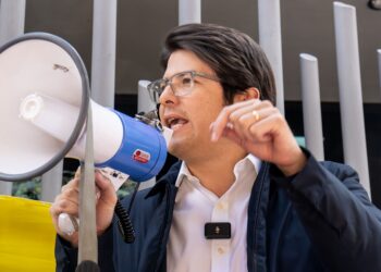 Miguel Uribe Turbay habla a través de un megáfono en un evento al aire libre, usando camisa blanca, chaqueta azul y gafas, gesticulando mientras se dirige a los asistentes.