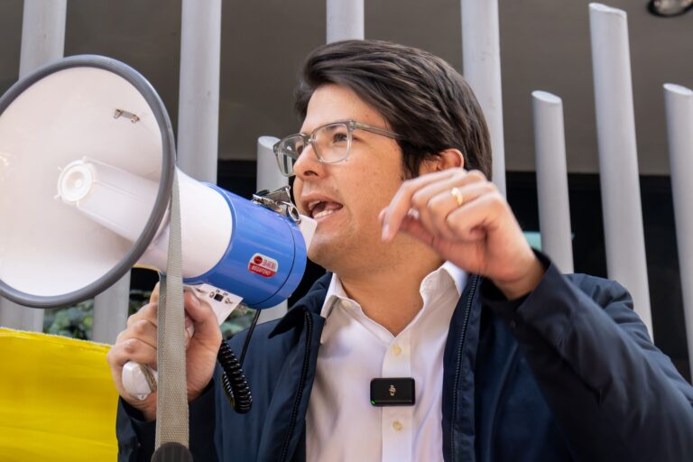 Miguel Uribe Turbay habla a través de un megáfono en un evento al aire libre, usando camisa blanca, chaqueta azul y gafas, gesticulando mientras se dirige a los asistentes.