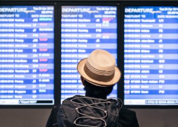 Persona con sombrero y mochila observa el tablero de información de vuelos de Aeroméxico en un aeropuerto.