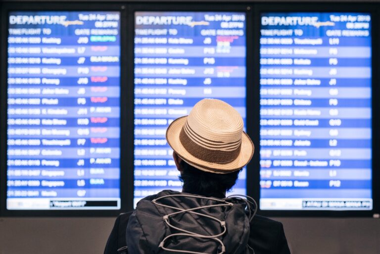 Persona con sombrero y mochila observa el tablero de información de vuelos de Aeroméxico en un aeropuerto.