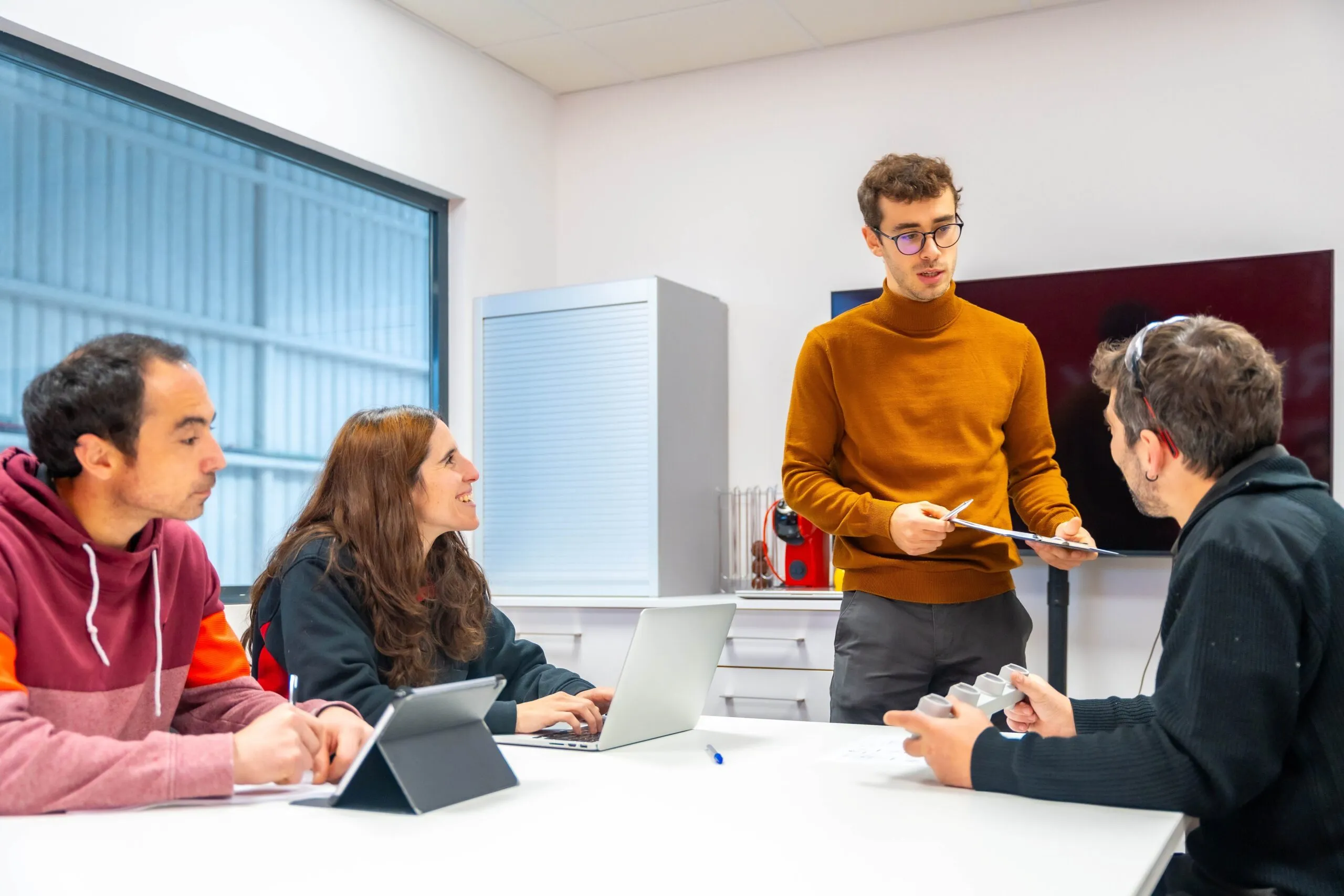 Grupo de cuatro personas participando en una reunión, con intercambio de ideas y uso de dispositivos electrónicos en una sala de trabajo iluminada.