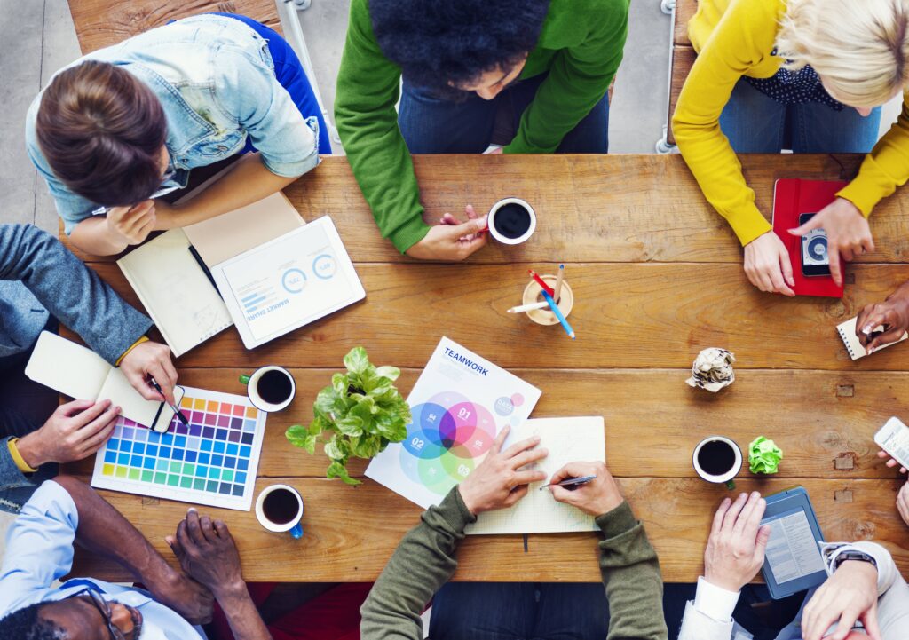 Vista aérea de un grupo diverso de personas trabajando en equipo alrededor de una mesa, revisando gráficos y paletas de color, representando la colaboración en la elaboración de políticas culturales en España.