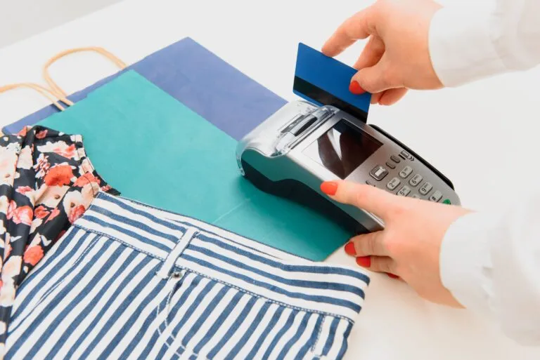 Manos deslizando tarjeta en terminal de pago junto a ropa y bolsas de compras, ilustrando cómo Gen Z impulsa la cultura del capricho que impacta finanzas personales.
