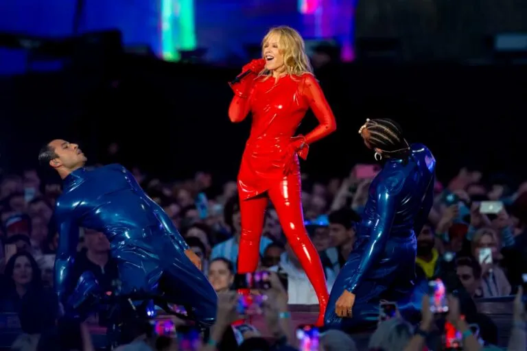 Kylie Minogue en concierto en la Ciudad de México durante el Tension Tour 2025, luciendo un traje rojo brillante junto a dos bailarines, frente a miles de fans en el Palacio de los Deportes.